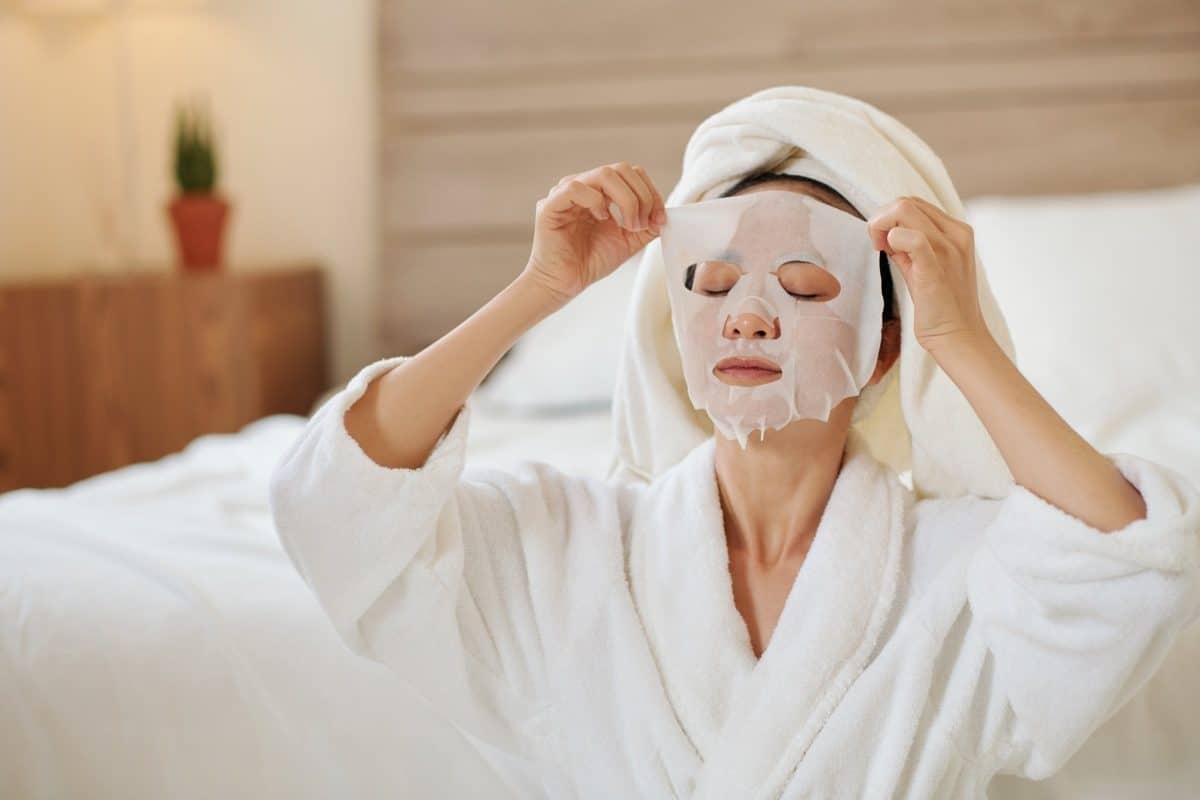 Woman placing face sheet mask on her face with eyes closed, dressed in a white gown and white towel wrapped on her head.